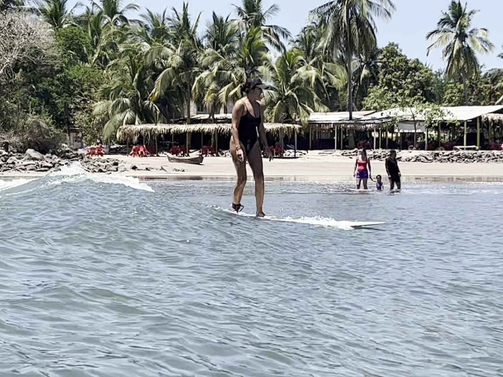 Alumna de pie sobre la tabla corriendo una ola, con las palapas y las palmeras al fondo