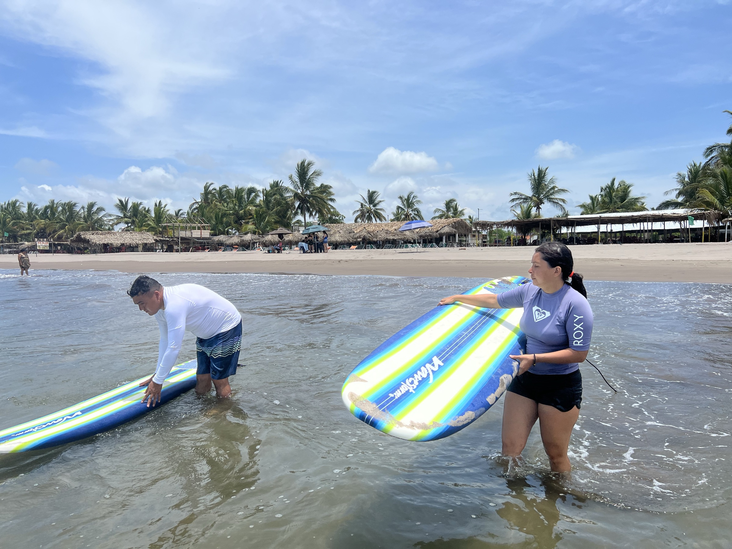 Instructor y alumna entrando al agua con sus tablas, con las palapas al fondo