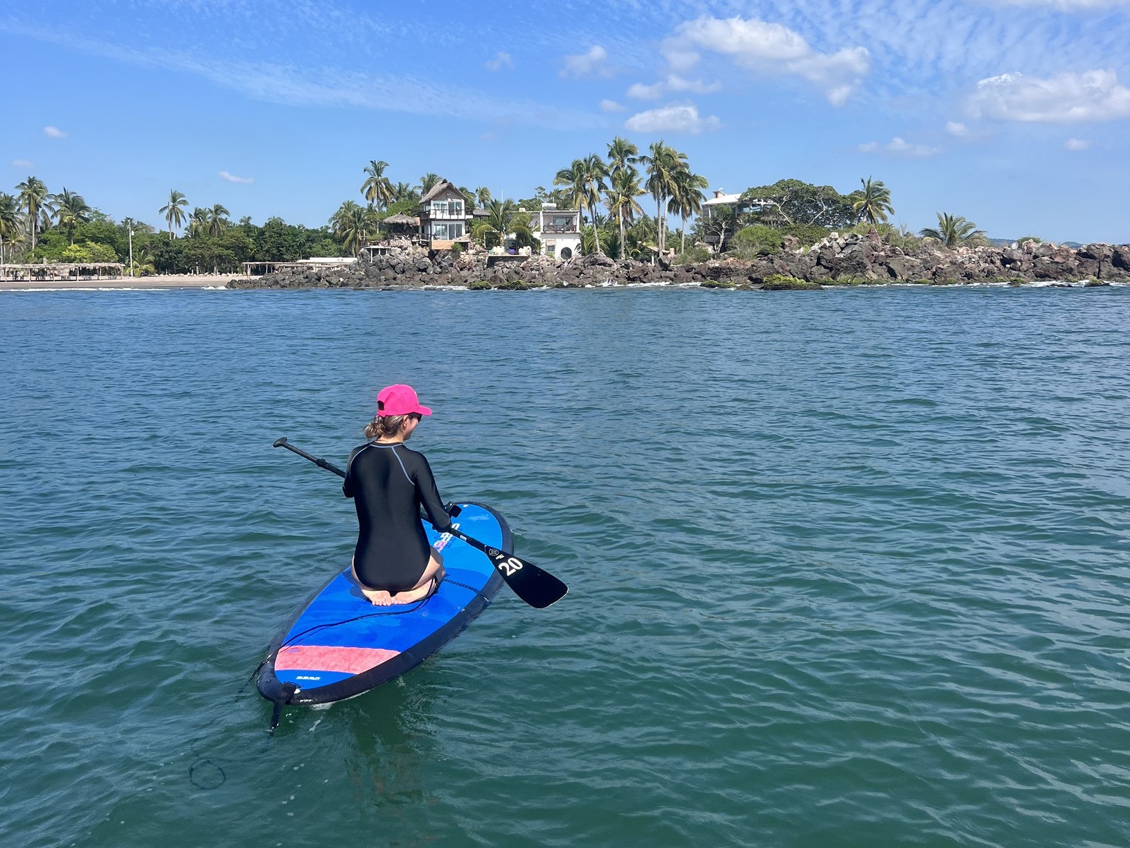 Alumna sobre un paddleboard en el agua plana de Las Islitas, con la isla y las palmeras al fondo
