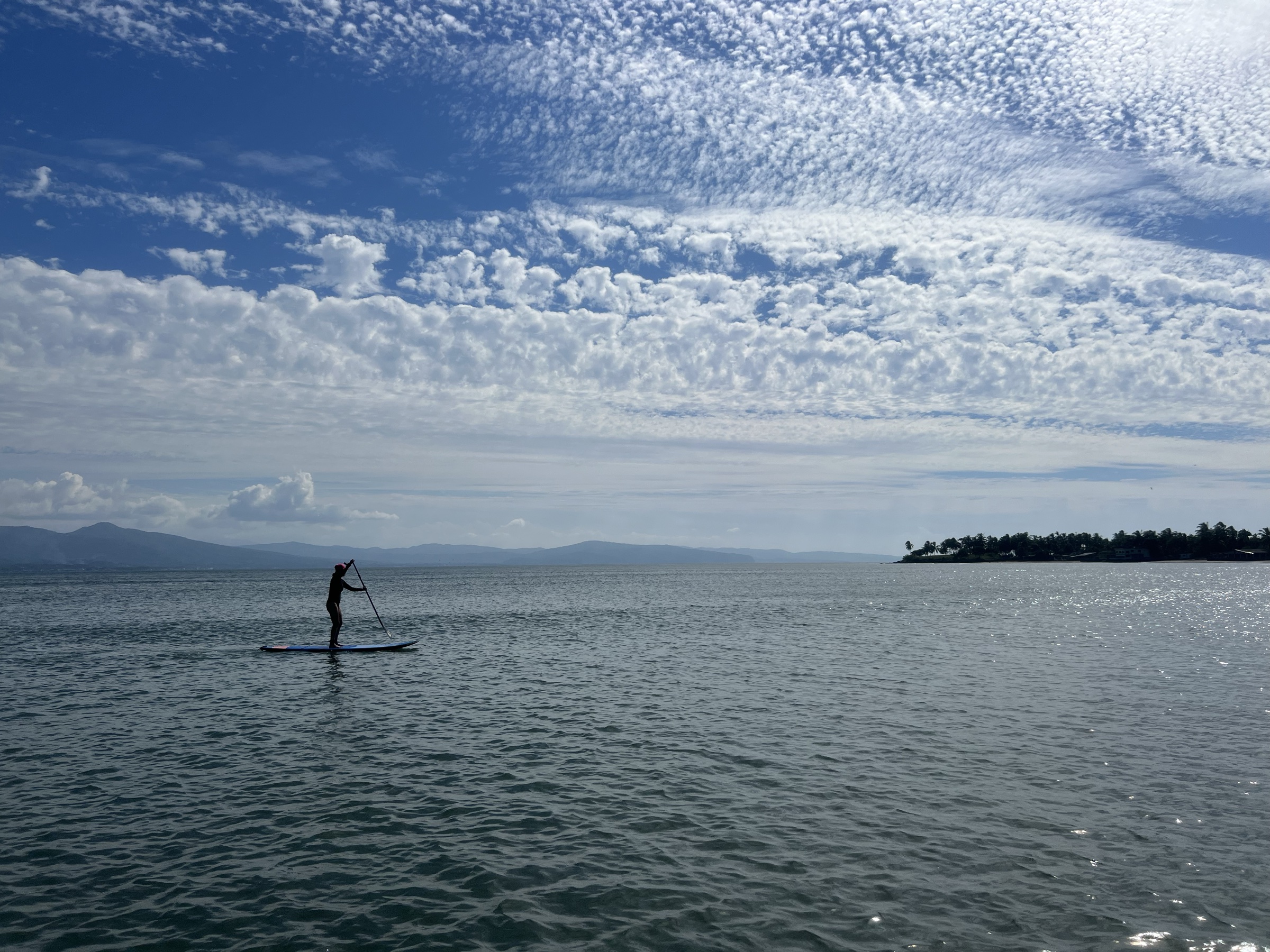 Persona remando de pie sobre un paddleboard en el agua plana de Las Islitas, con la sierra y una isla al fondo