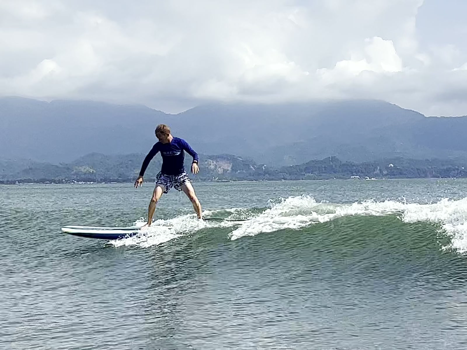 Alumno de pie sobre la tabla corriendo una ola, con la bahía y la sierra de San Blas al fondo