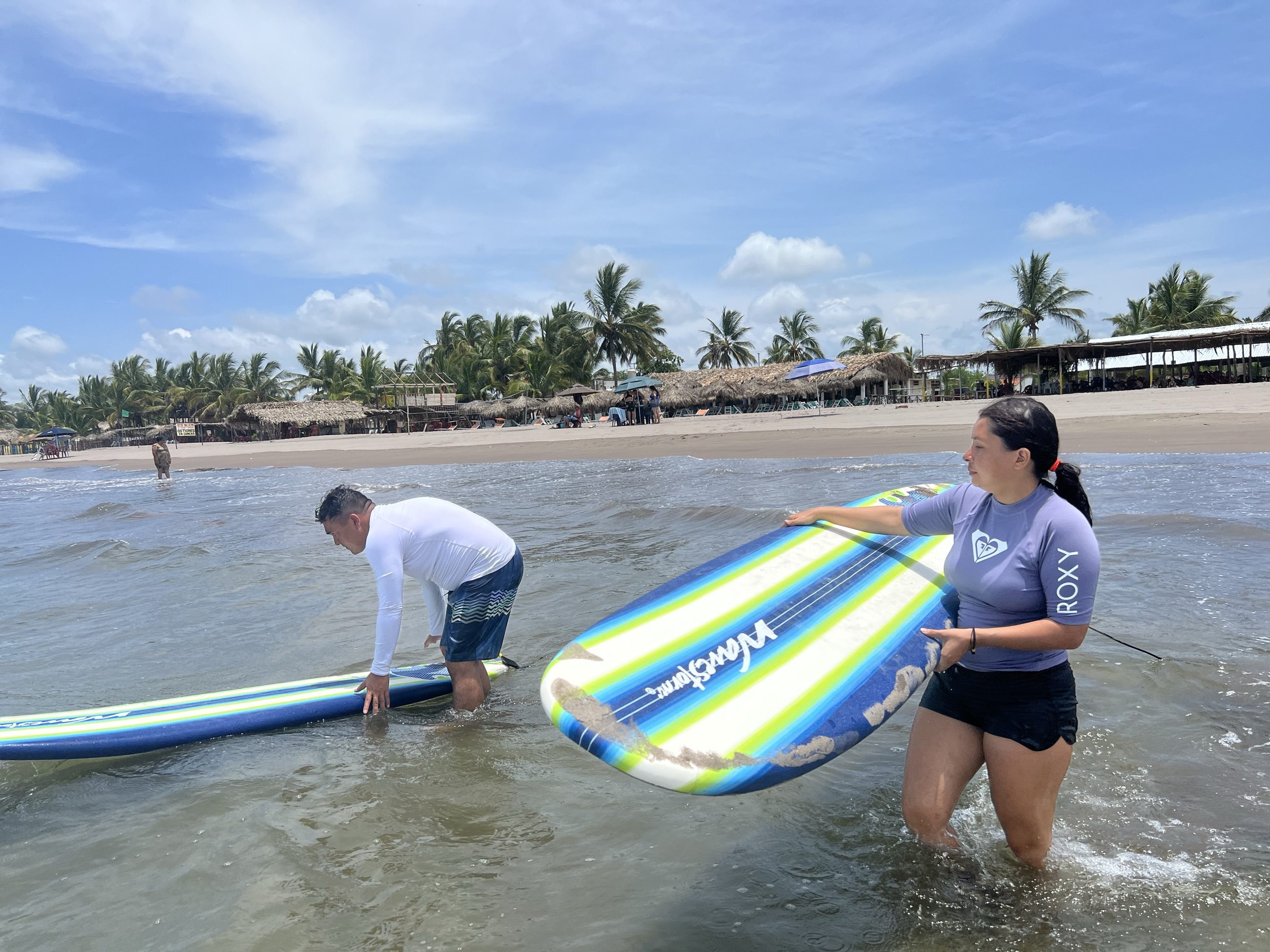 Instructor y alumna metiendo sus tablas al agua al empezar la clase, con las palapas de la playa al fondo