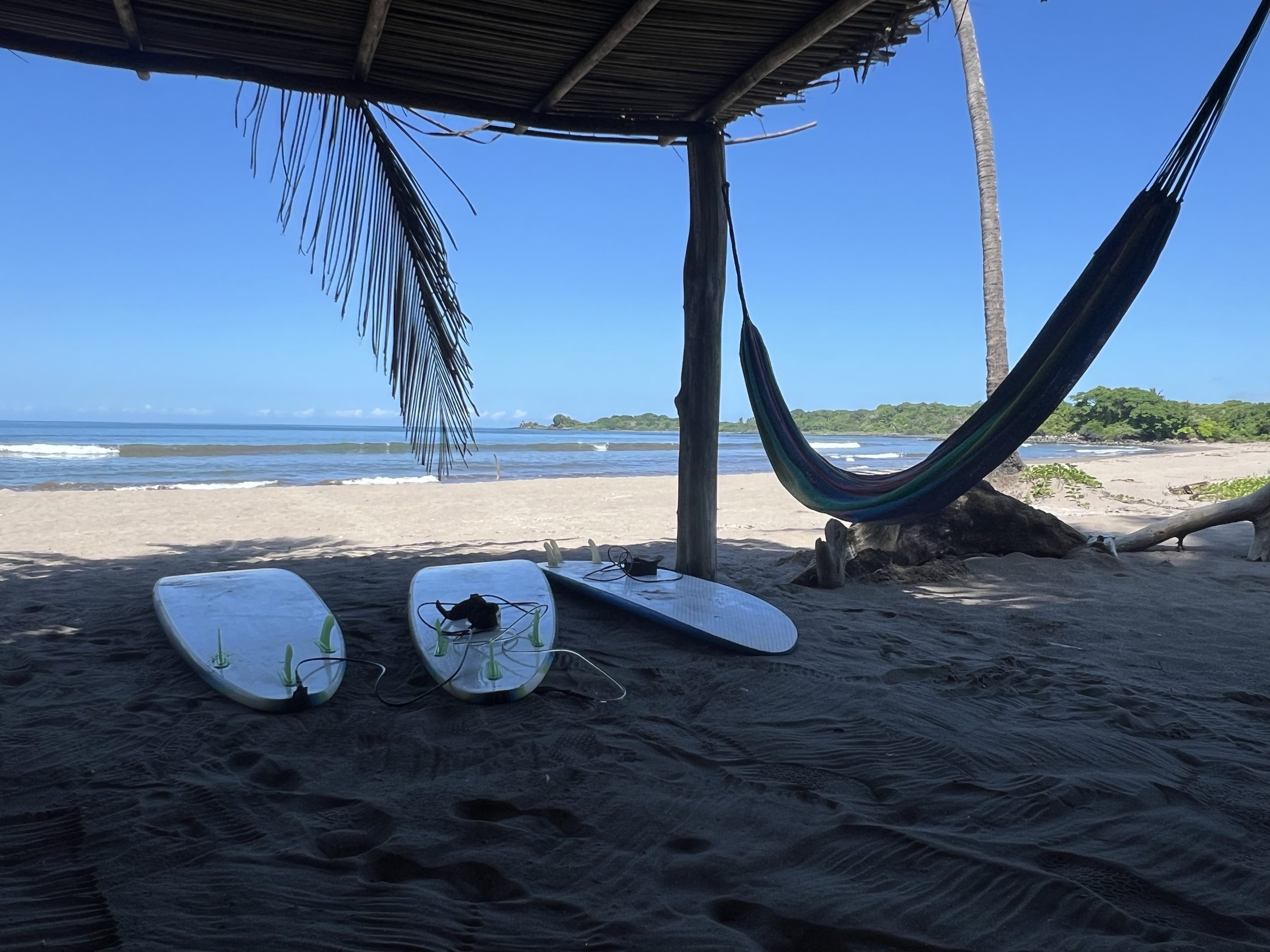 Palapa frente a la playa con tablas sobre la arena y una hamaca colgada junto a la palmera