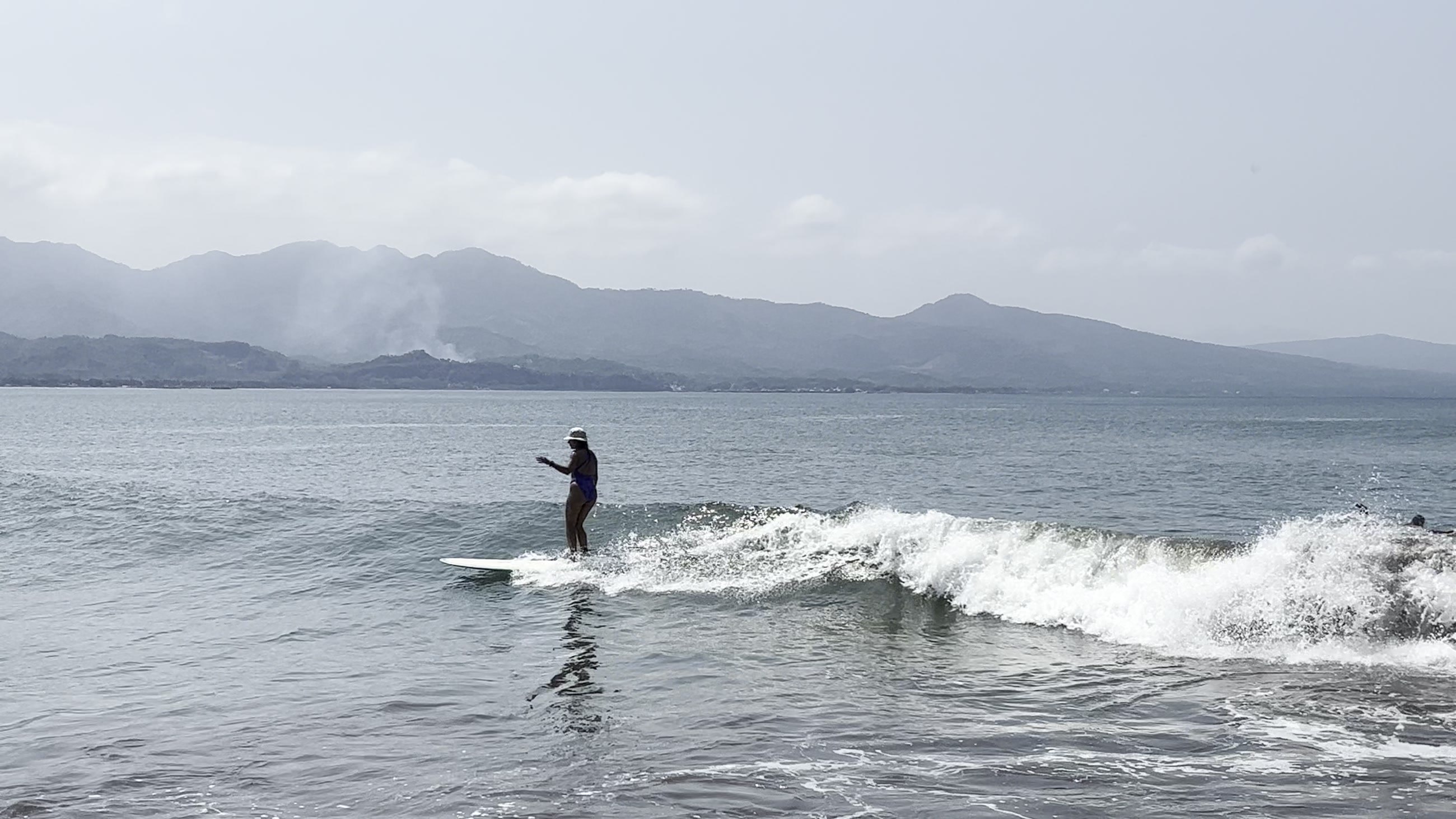 Surfista de pie sobre una tabla larga recorriendo una ola pequeña y bien formada, con la sierra al fondo