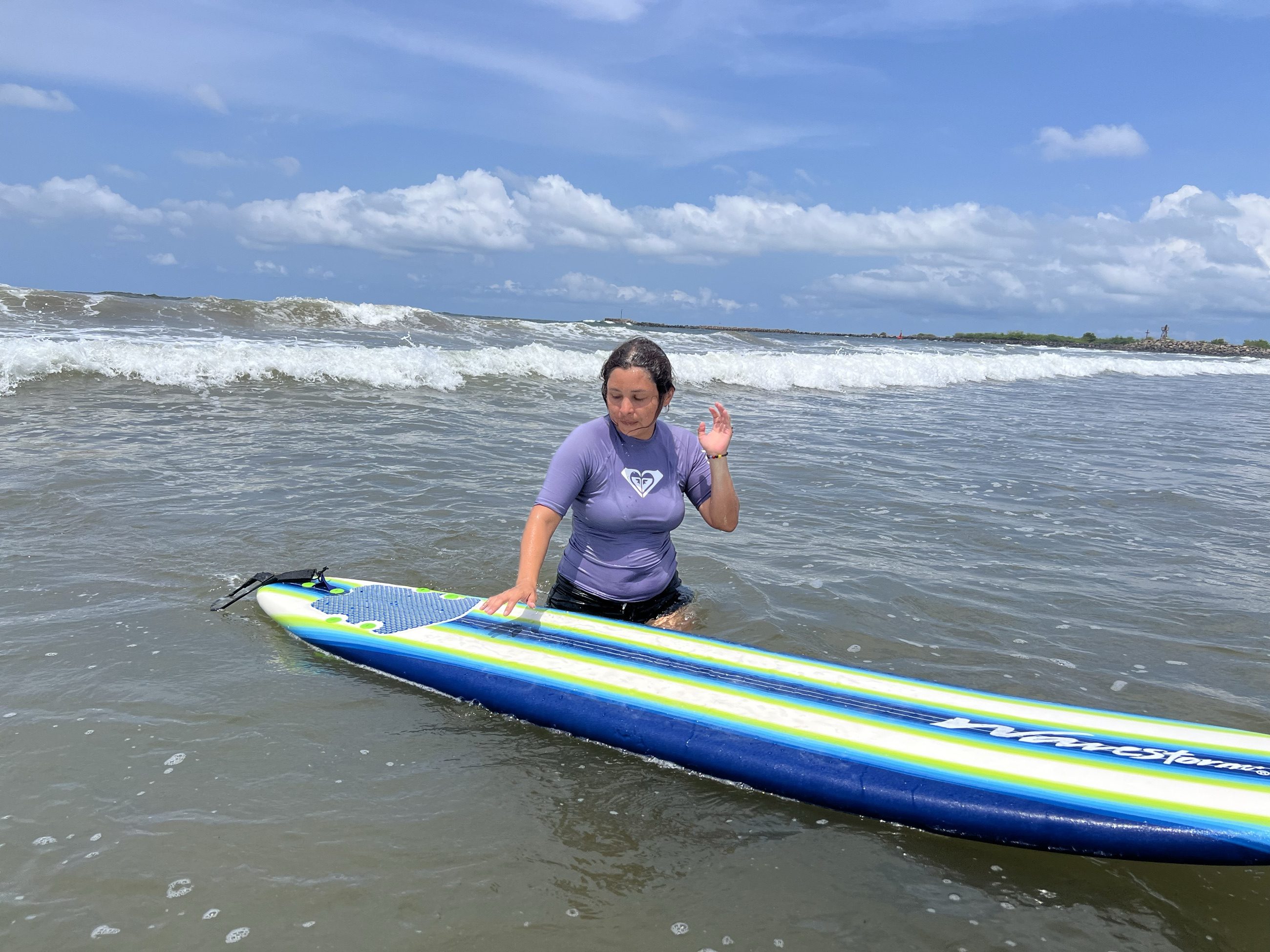 Alumna con rashguard morado junto a su tabla, sonriendo y saludando